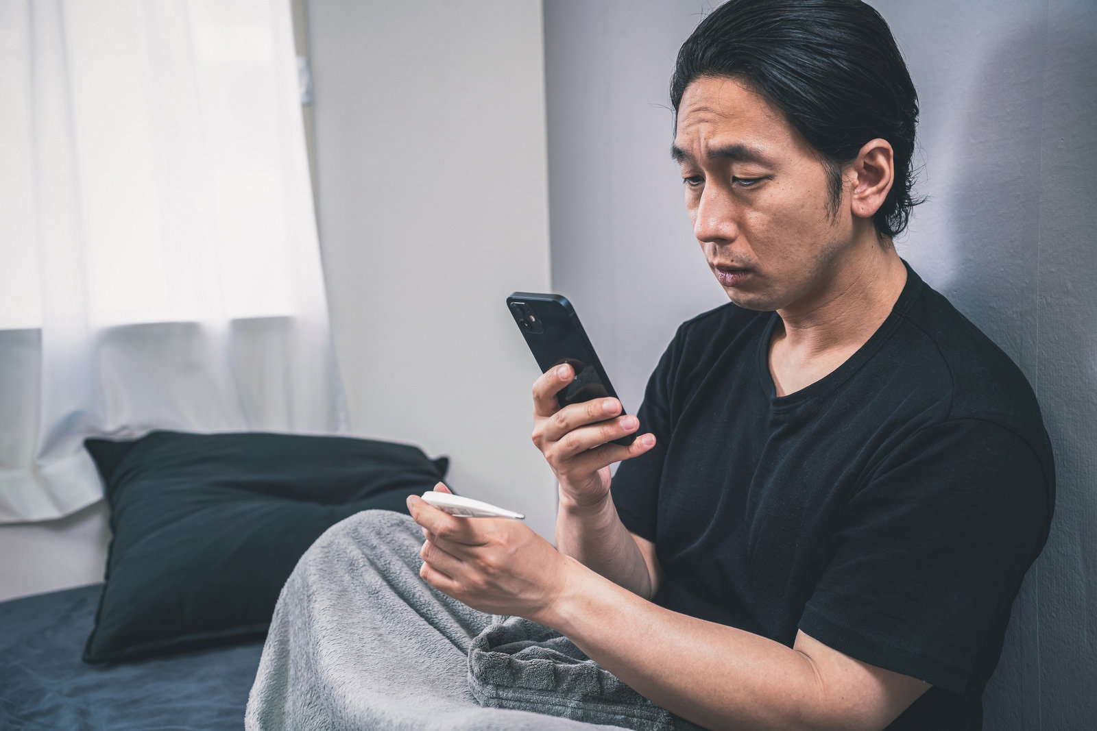 黒いTシャツを着た男性が体温計を持ちながらスマートフォンを見ている様子［モデル：大川竜弥］