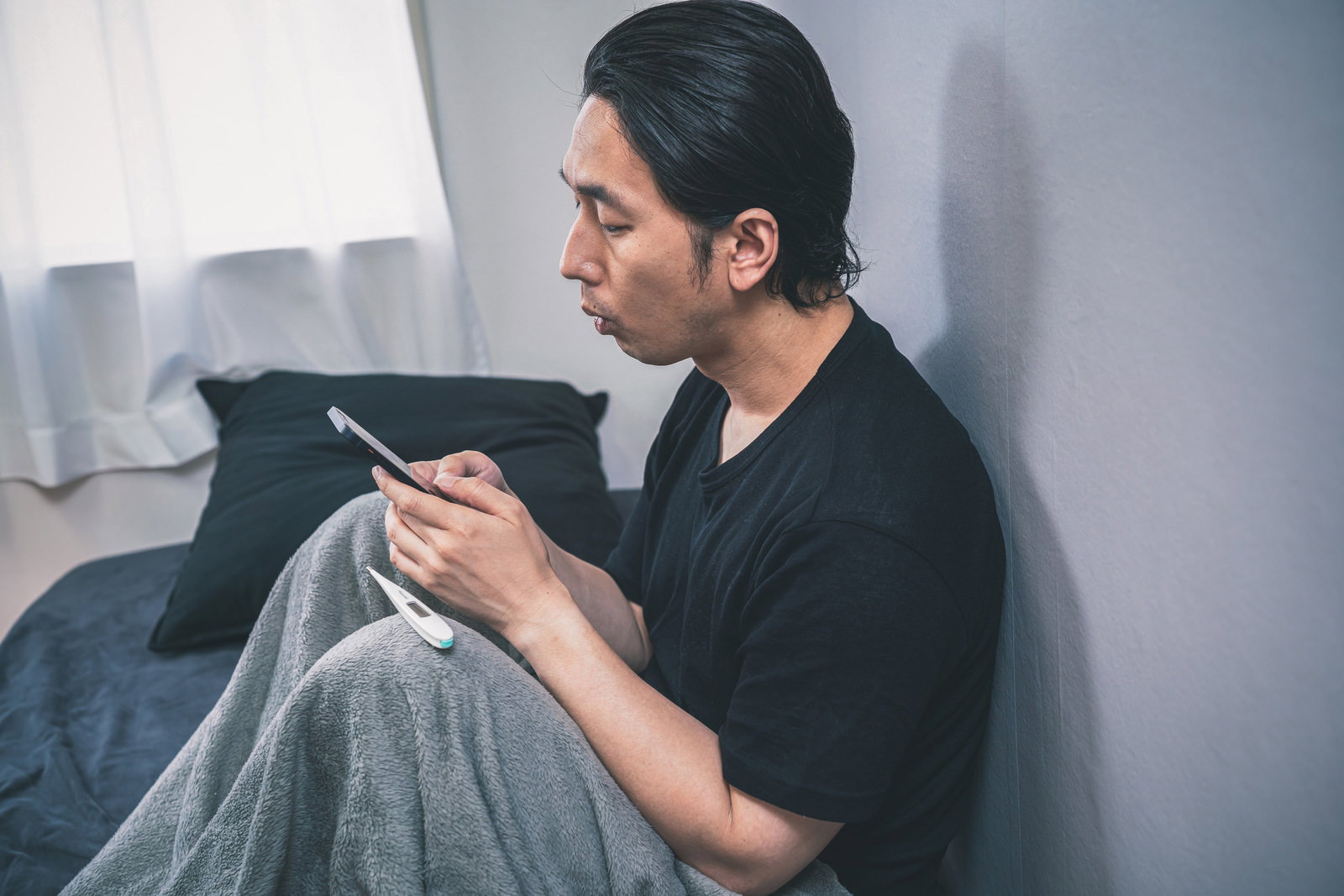 ベッドで横になりながらスマートフォンで連絡をする体調不良の男性［モデル：大川竜弥］