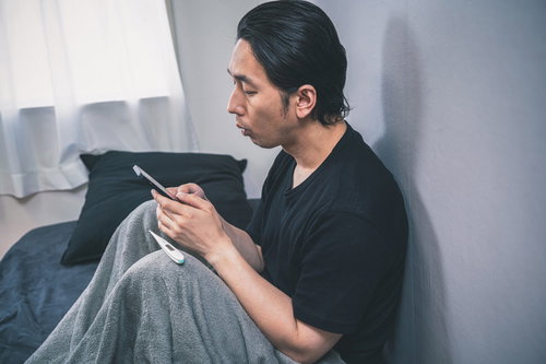 体調不良で職場に連絡をするベッドの男性、体温計を持つ