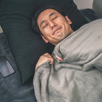 枕元にスマホと体温計を置いて就寝する男性の写真