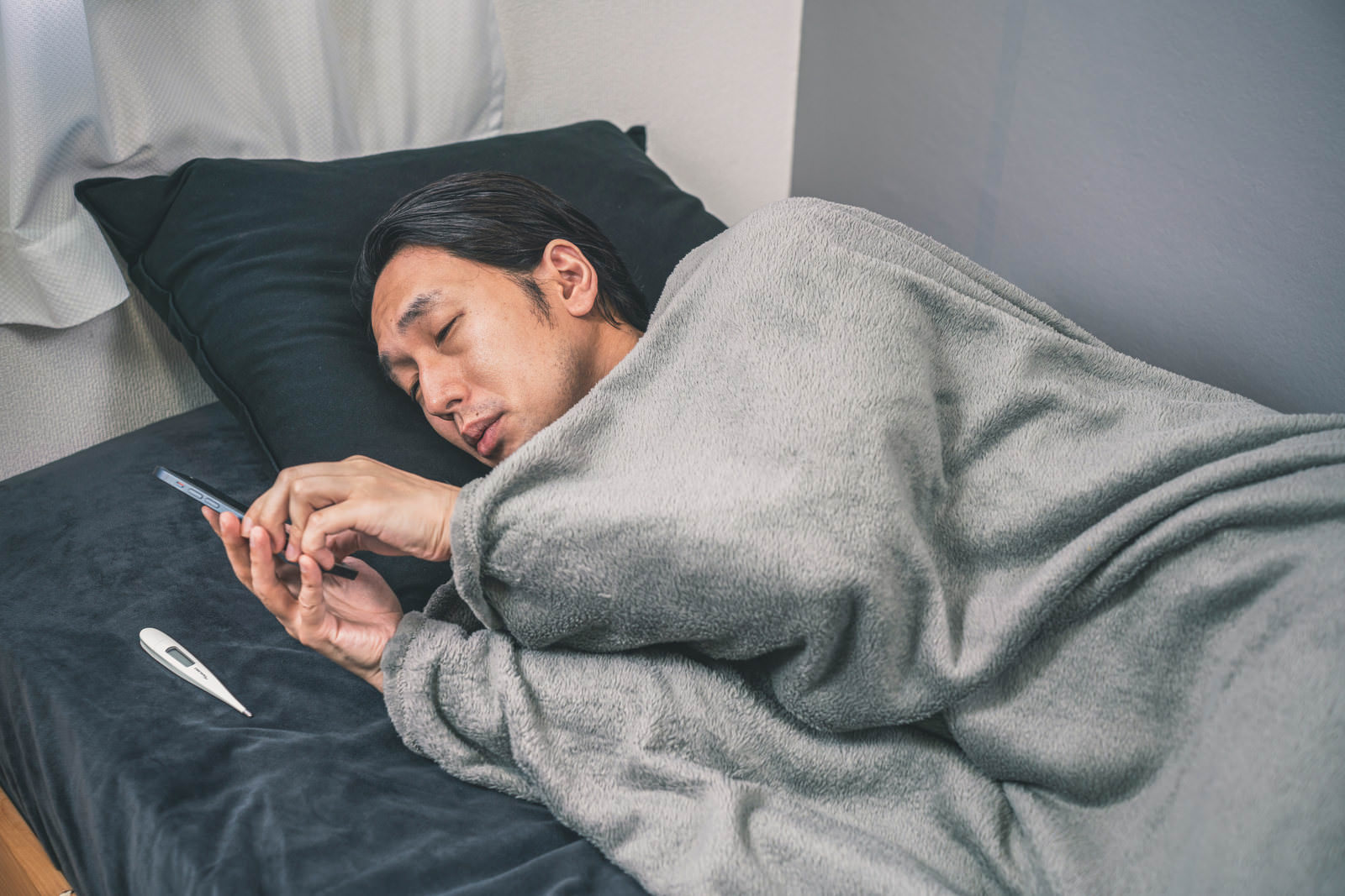 ベッドでスマートフォンを見る男性と枕元の体温計［モデル：大川竜弥］