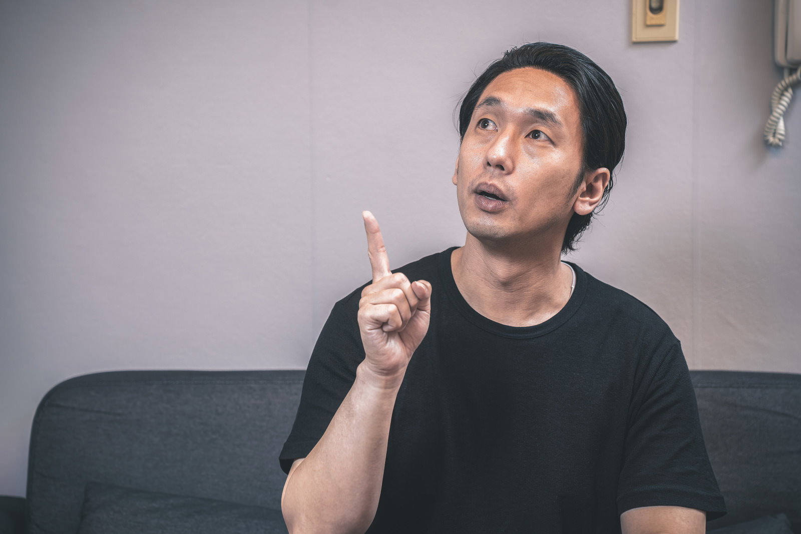 黒いTシャツを着た男性がソファに座って天井を指差している