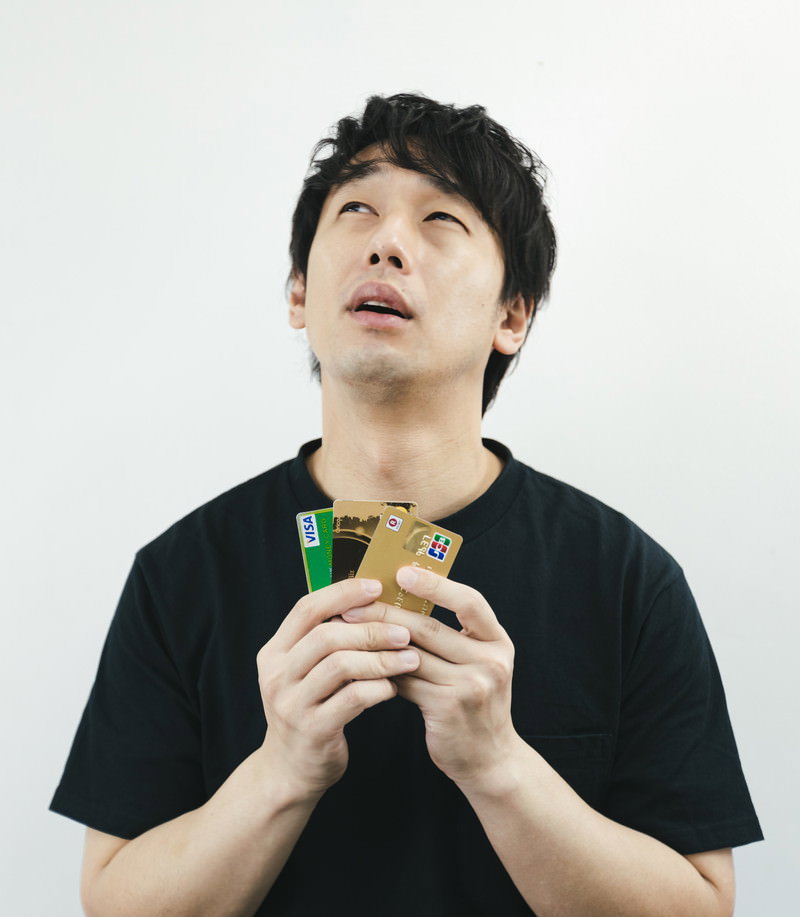 両手でクレジットカードを持ち上を見上げる黒いTシャツの男性［モデル：大川竜弥］