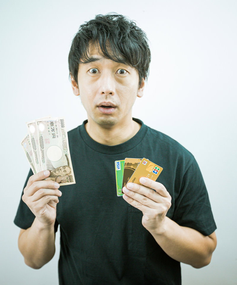 現金とクレジットカードを両手に持ち迷う表情の男性［モデル：大川竜弥］
