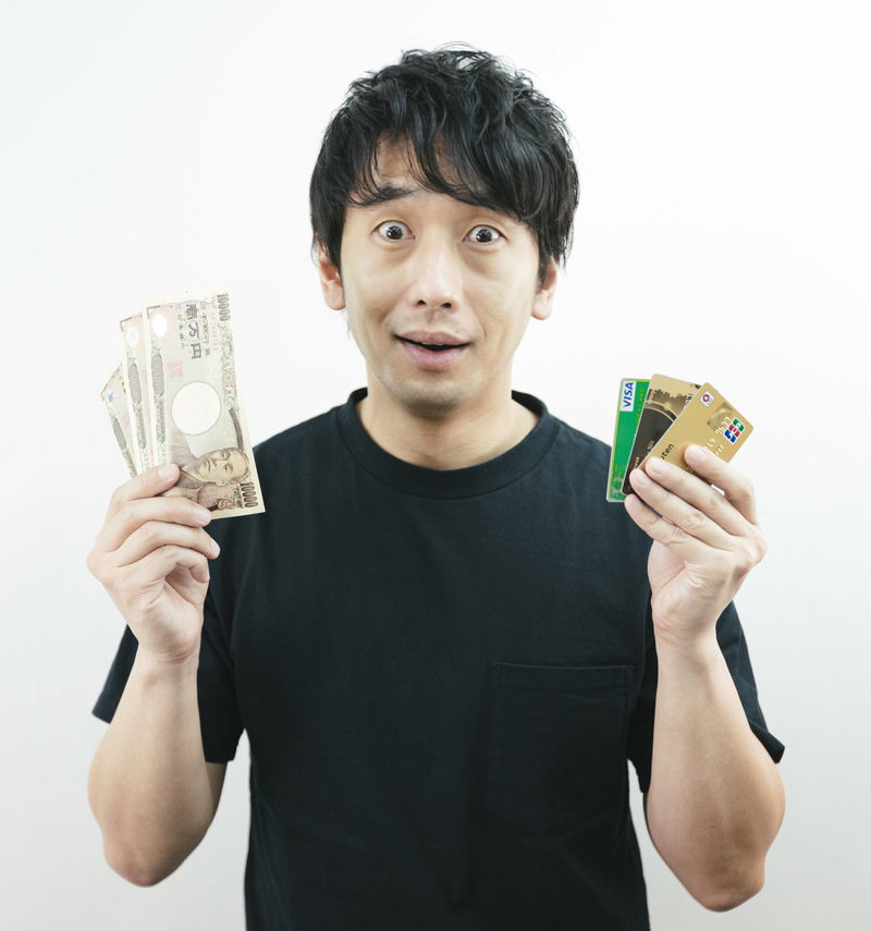 現金とクレジットカードを両手に持ち迷っている表情の男性［モデル：大川竜弥］