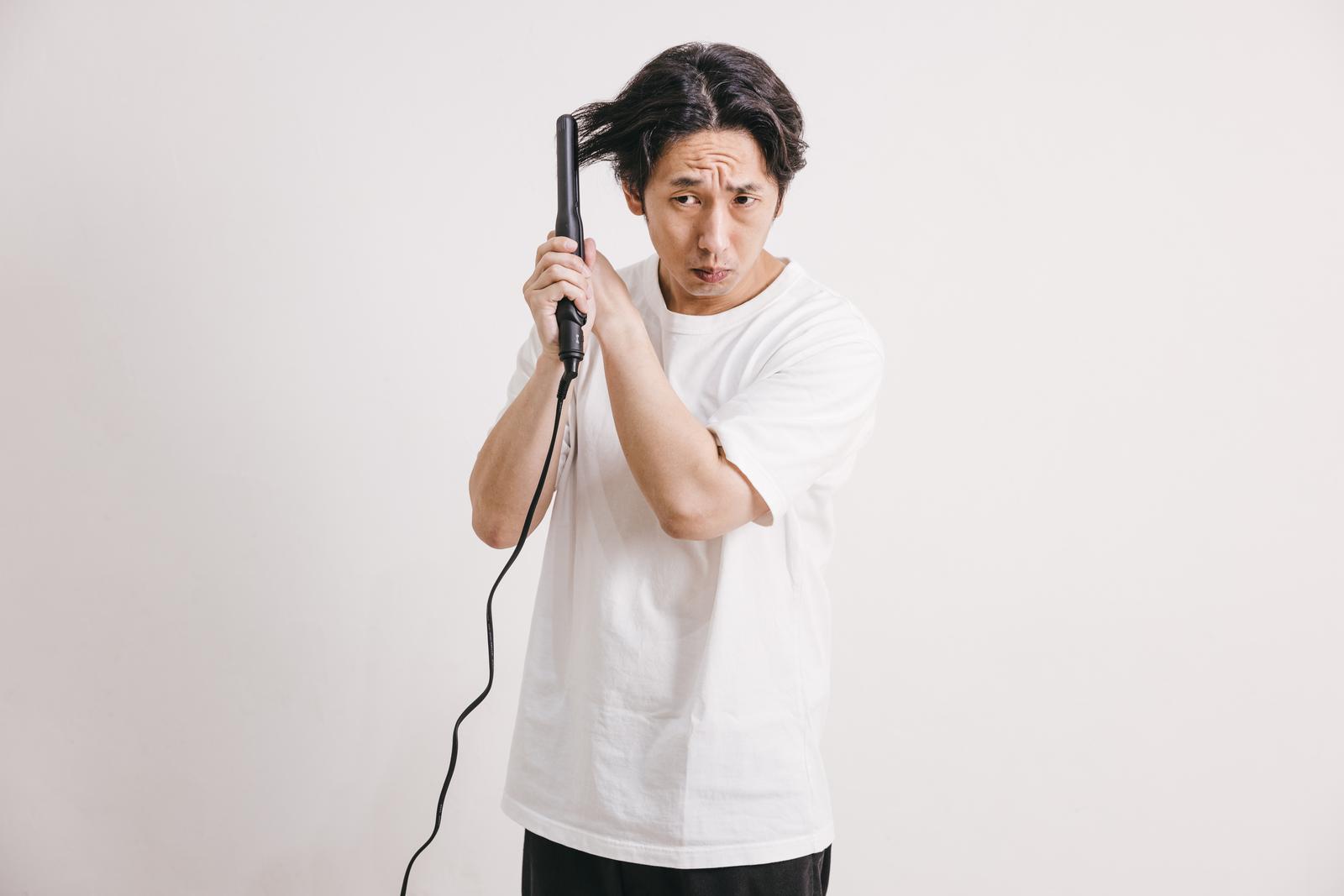 黒いヘアアイロンを手に持ち不安そうな表情を見せる白いTシャツの男性［モデル：大川竜弥］