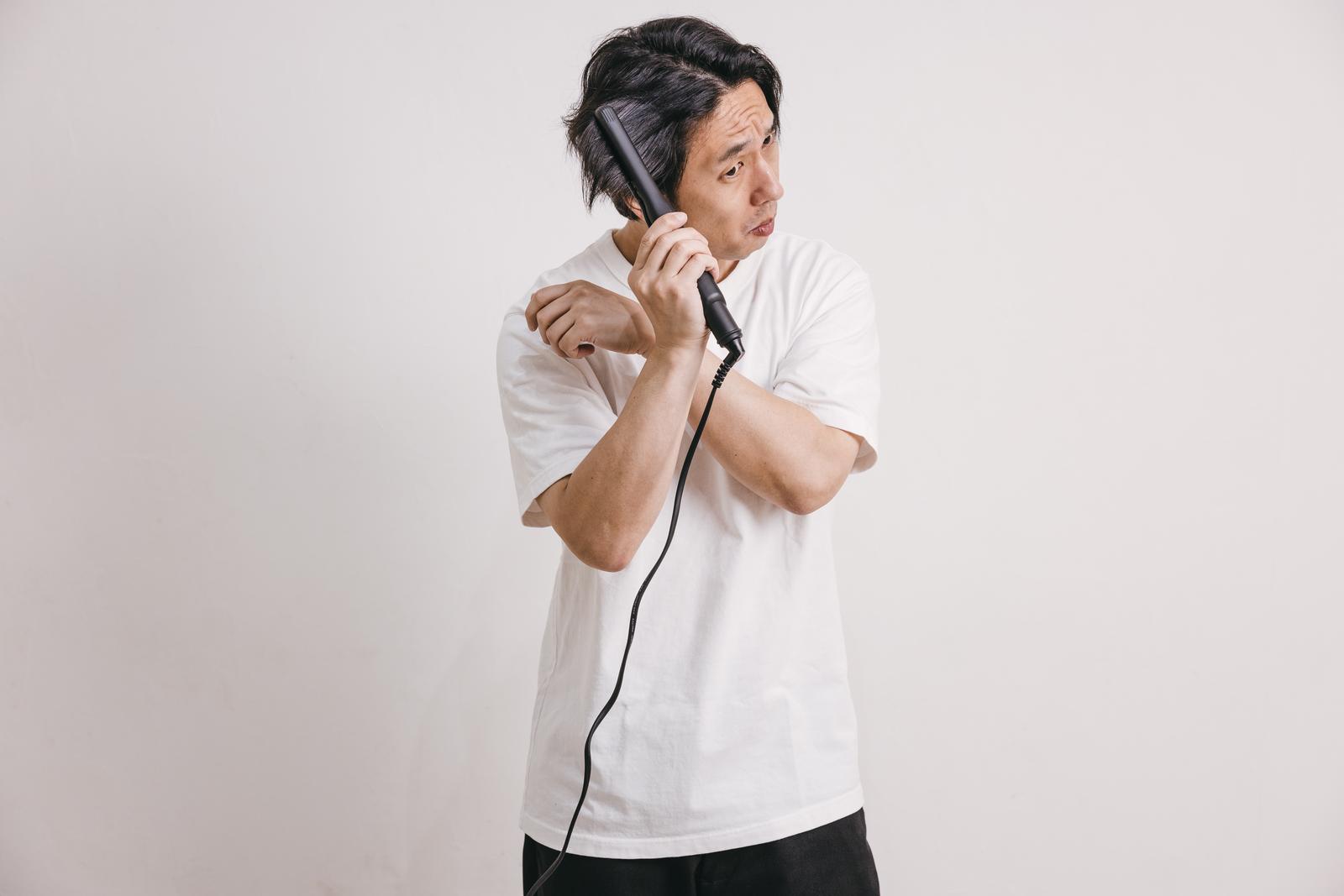白いTシャツの男性が黒いヘアアイロンを右手で持ち髪に当てている［モデル：大川竜弥］
