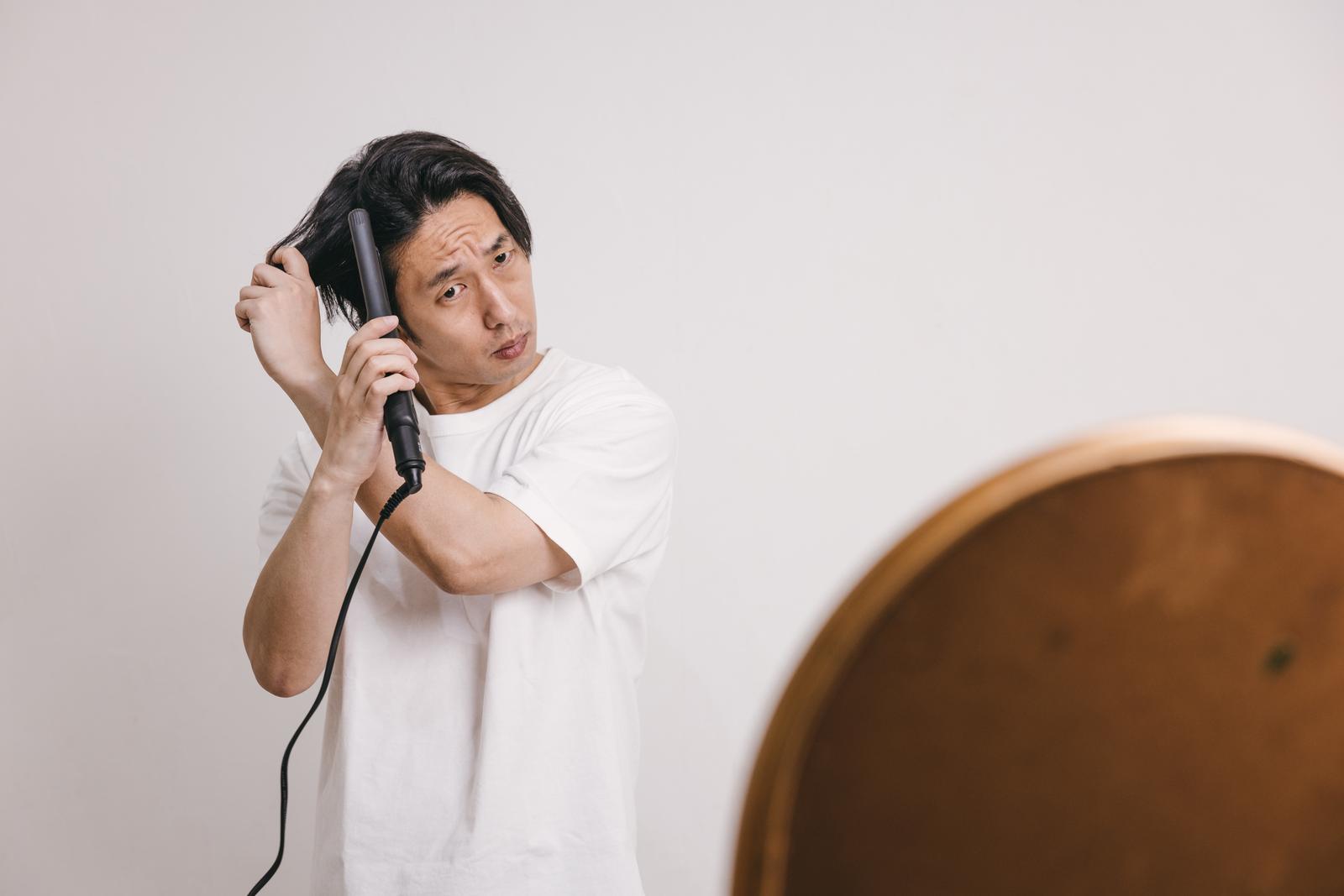 鏡を見ながらヘアアイロンで髪をセットしている白いTシャツの男性［モデル：大川竜弥］