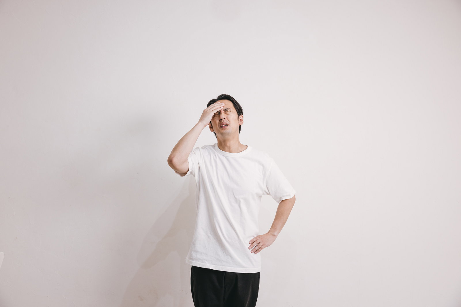 白いTシャツの男性が頭痛で手を頭に当てて困った表情をしている［モデル：大川竜弥］