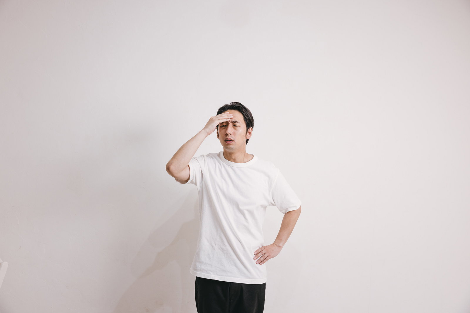 額に手を当てて頭痛に悩む白いTシャツの男性［モデル：大川竜弥］