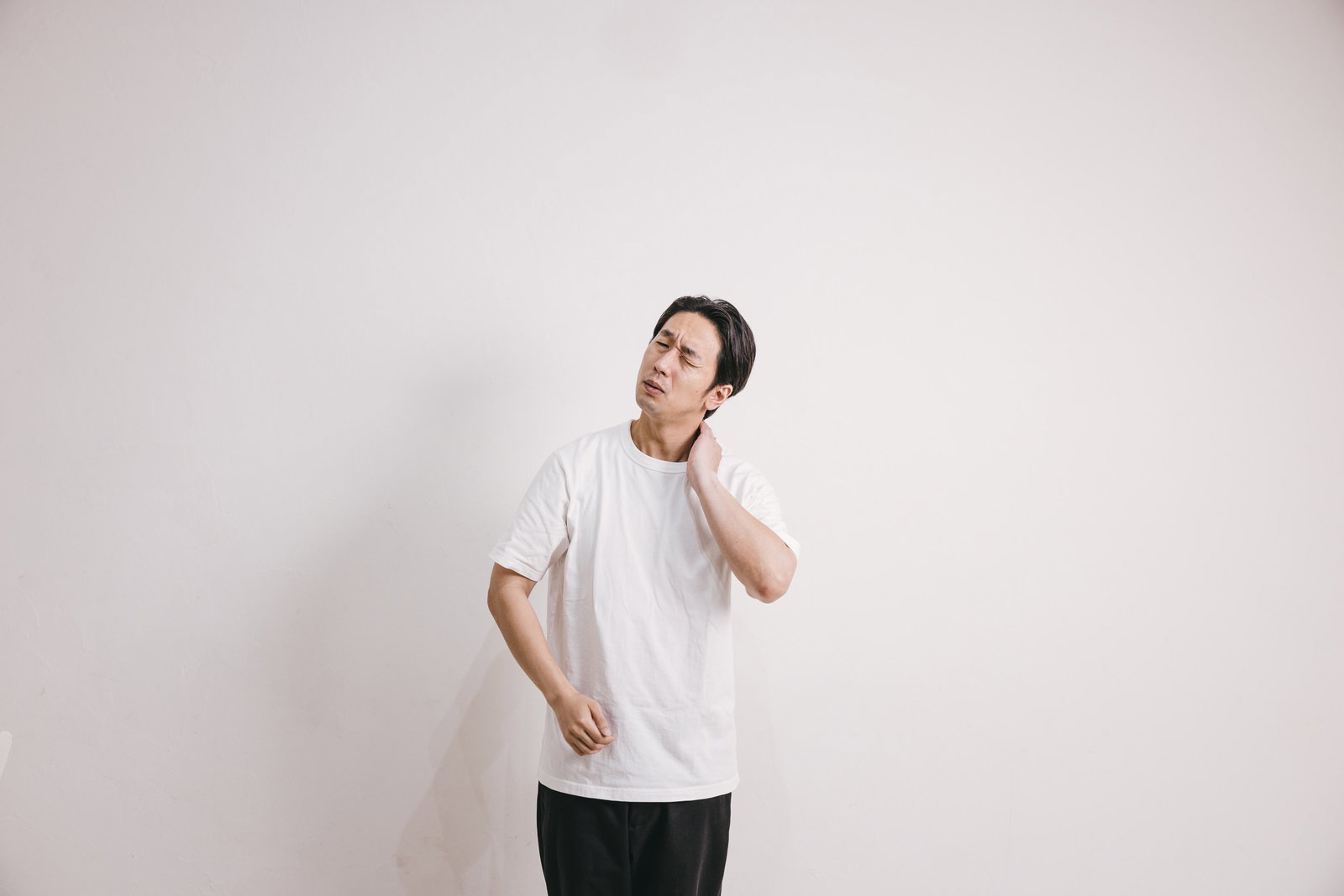 首の後ろに手を当てて痛そうな表情をする白いTシャツの男性［モデル：大川竜弥］