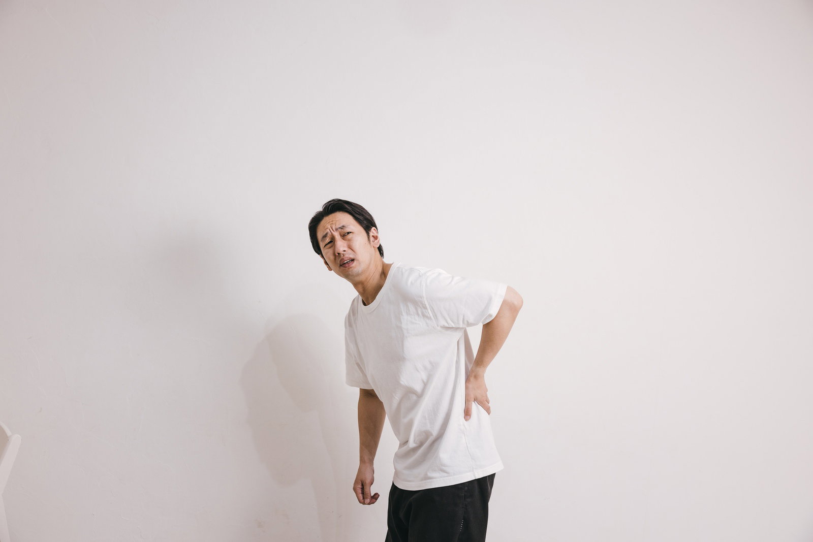 腰を押さえて痛がる表情の白いTシャツを着た男性［モデル：大川竜弥］