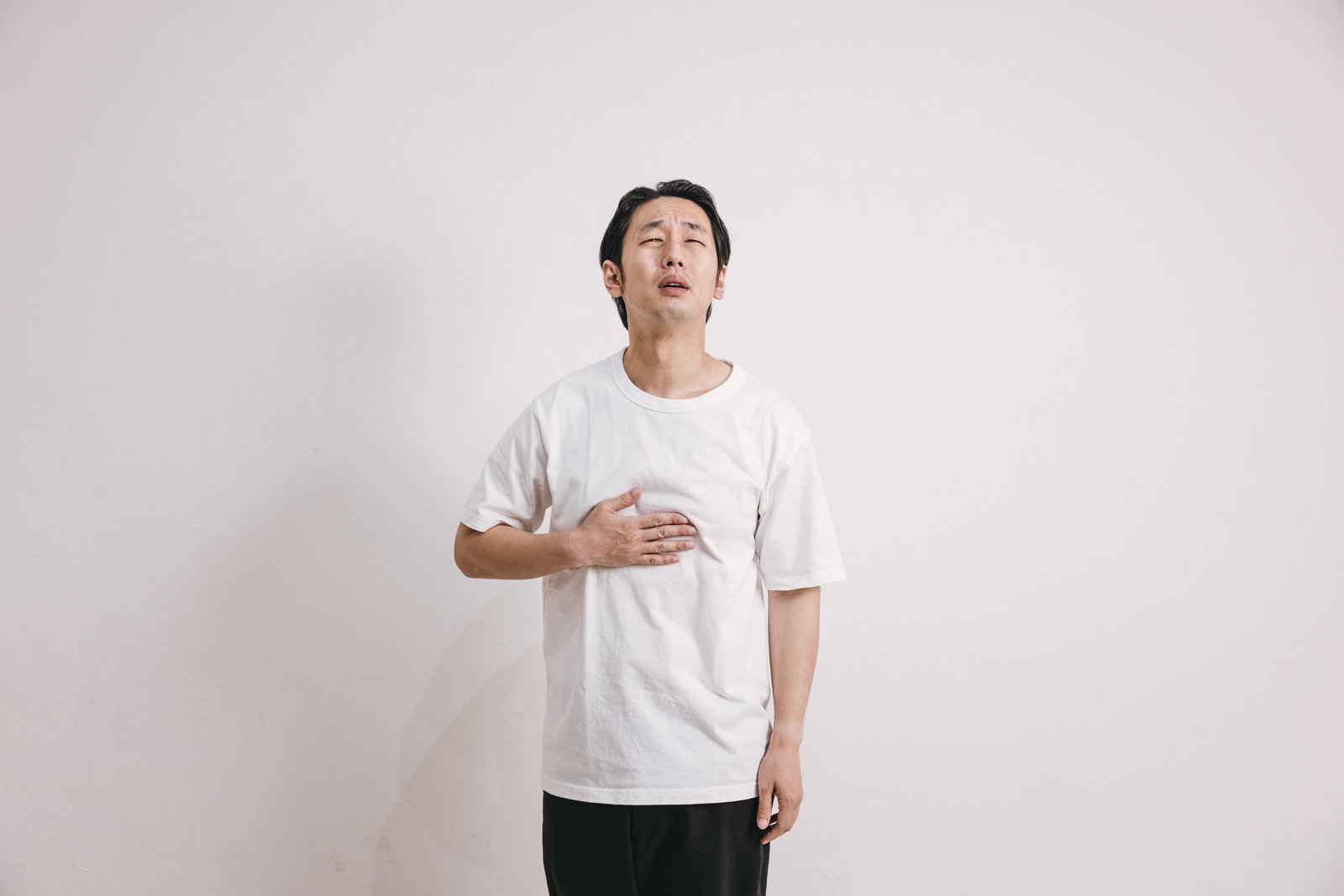 みぞおちに手を当てて痛みを訴える白いTシャツの男性［モデル：大川竜弥］