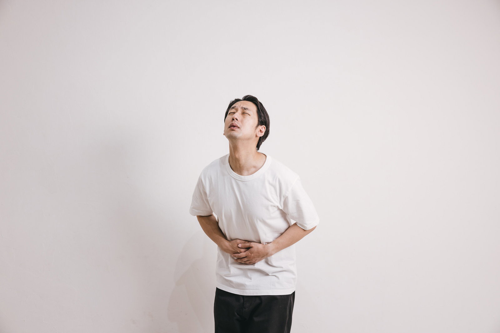 胃腸炎で腹部を押さえて苦しそうにする白いTシャツの男性［モデル：大川竜弥］