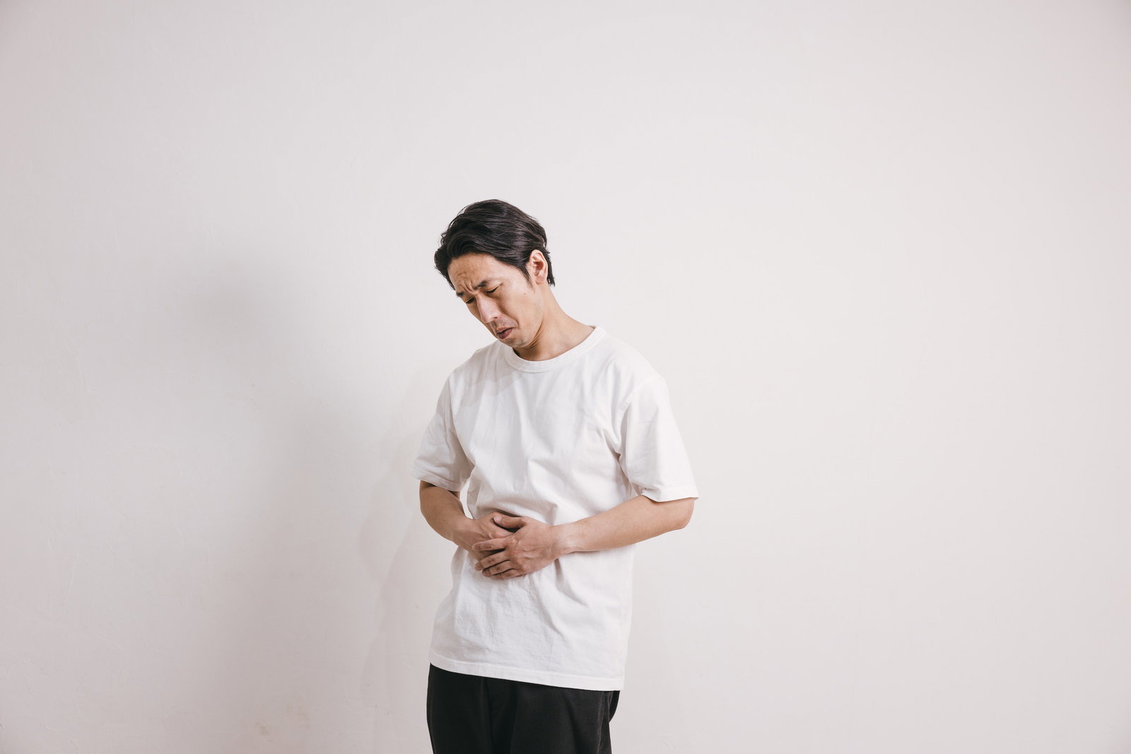 白いTシャツを着た男性がストレスで胃痛を訴えている様子［モデル：大川竜弥］
