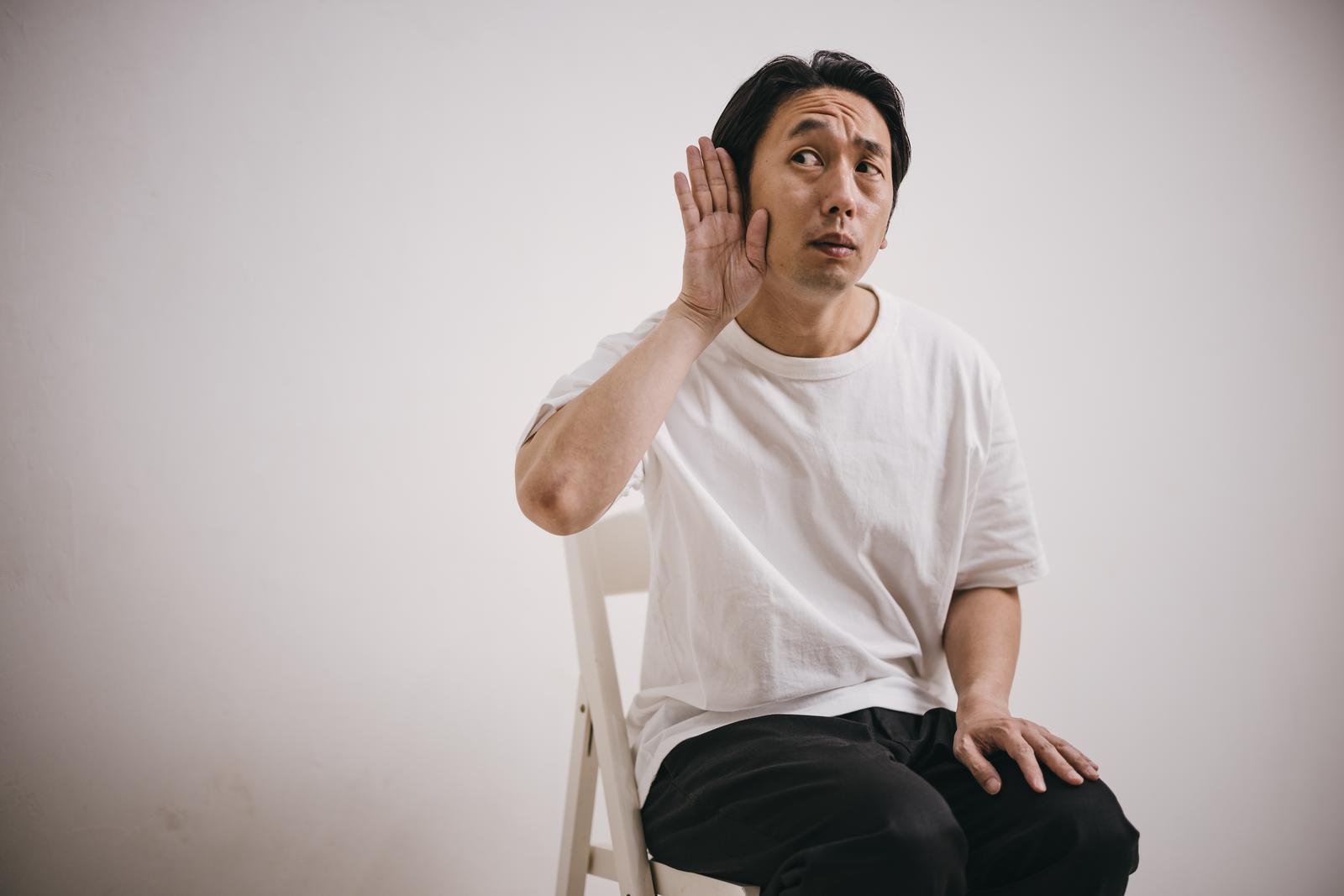 白いシャツの男性が手を耳に当てて聞き耳を立てるポーズ［モデル：大川竜弥］