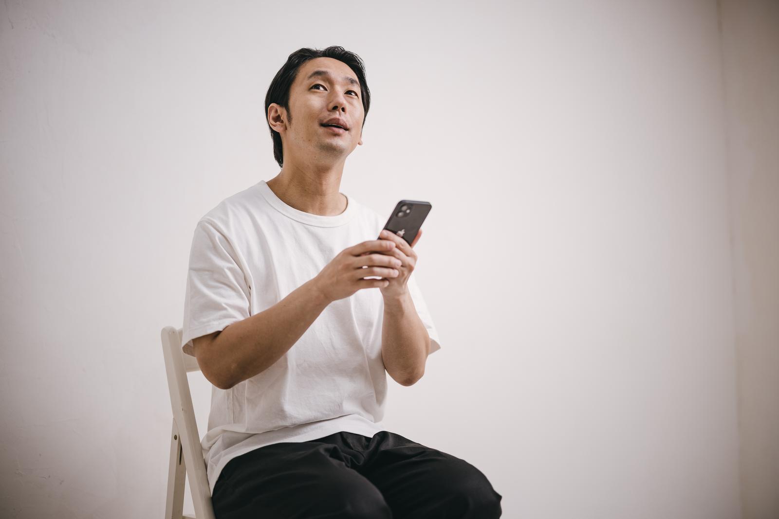 白いTシャツを着てスマホを持ち上を見上げる座っている男性［モデル：大川竜弥］