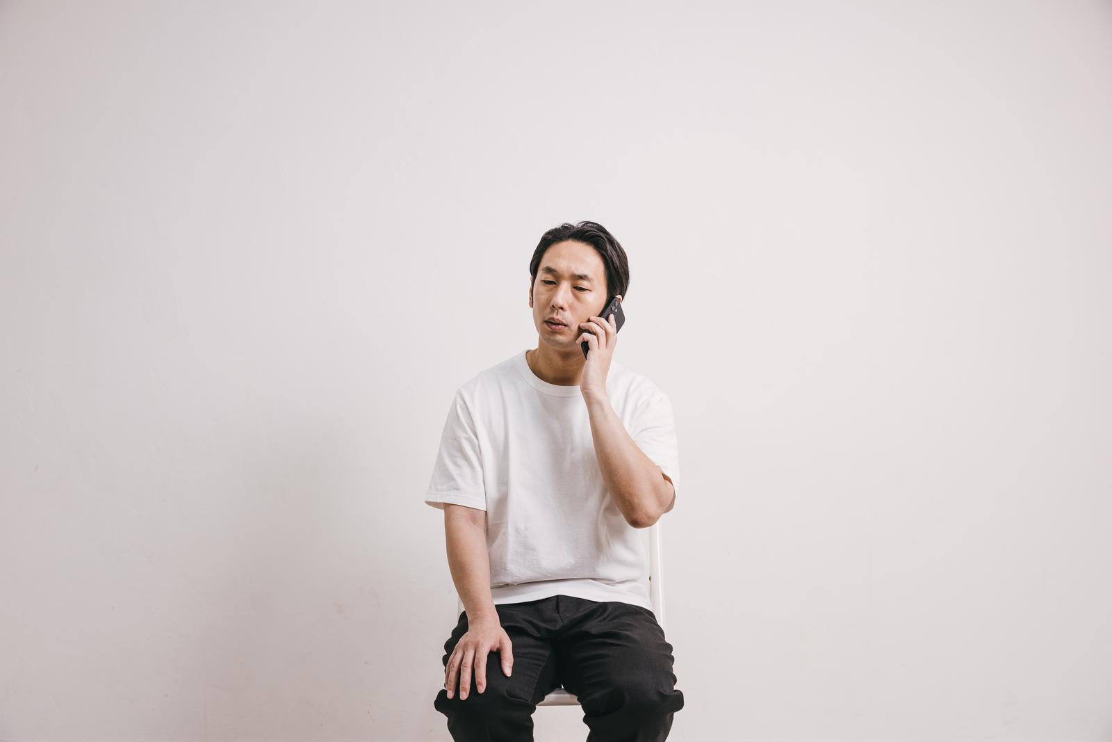 白い壁の前で電話をかけながら手を顔に当てて困った表情をする男性［モデル：大川竜弥］