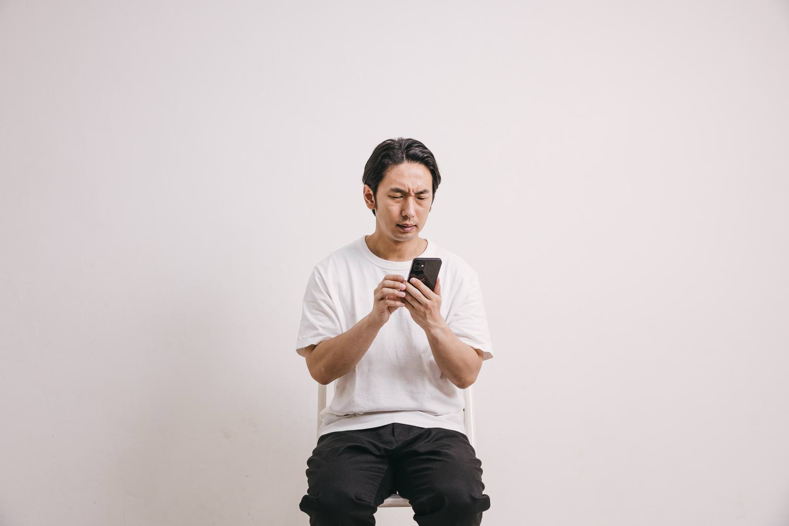 白いTシャツを着てスマートフォンを持ち、眉をひそめて怒った表情の男性が座っている［モデル：大川竜弥］