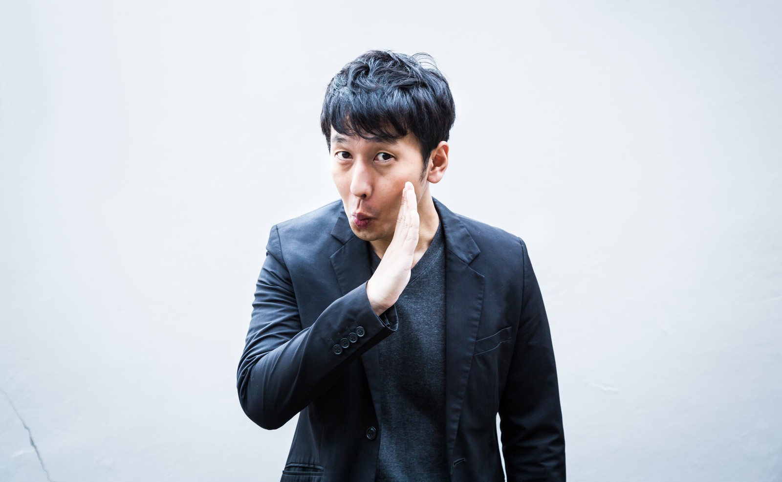片手を頬に当てて照れる黒スーツの短髪男性［モデル：大川竜弥］