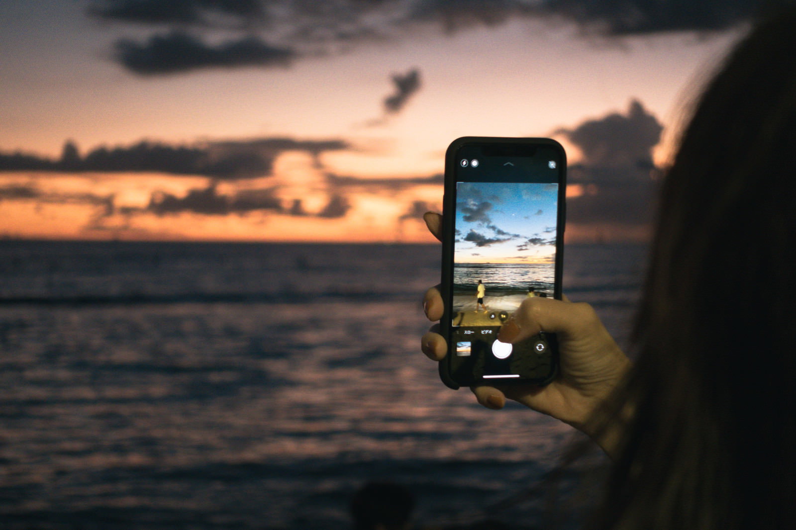 夕焼けの海でスマートフォンで波打ち際を撮影している人のシルエット