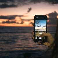 スマホで夕焼けと波打ち際を撮影する人のシルエットの写真