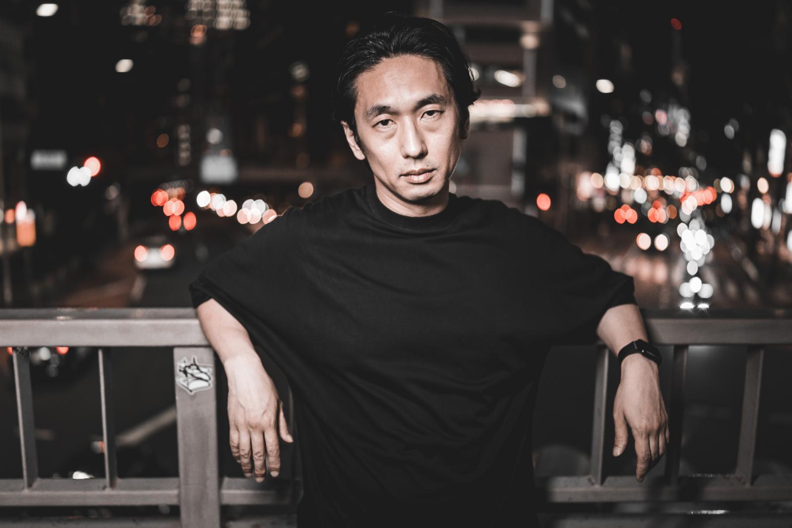 夜の橋で欄干に寄りかかり決めポーズを取る黒いTシャツの男性［モデル：大川竜弥］