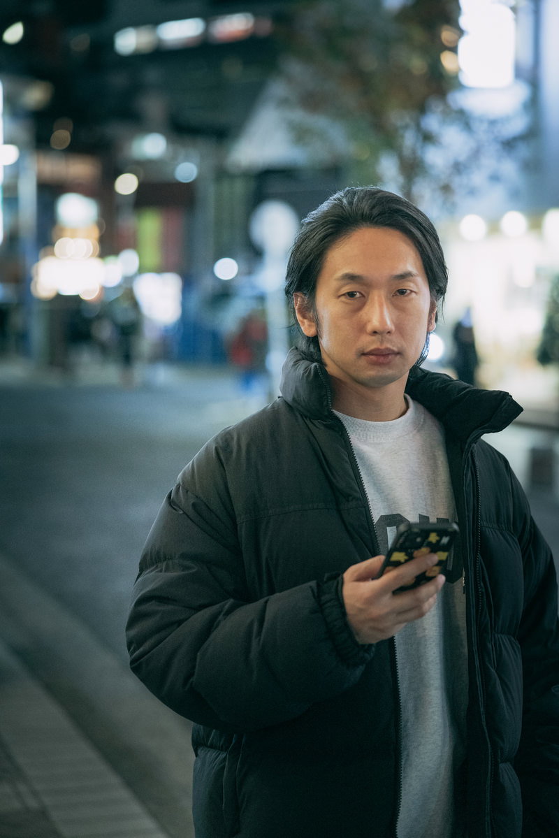 夜の繁華街でスマートフォンを見つめる黒いダウンジャケットを着た男性［モデル：大川竜弥］