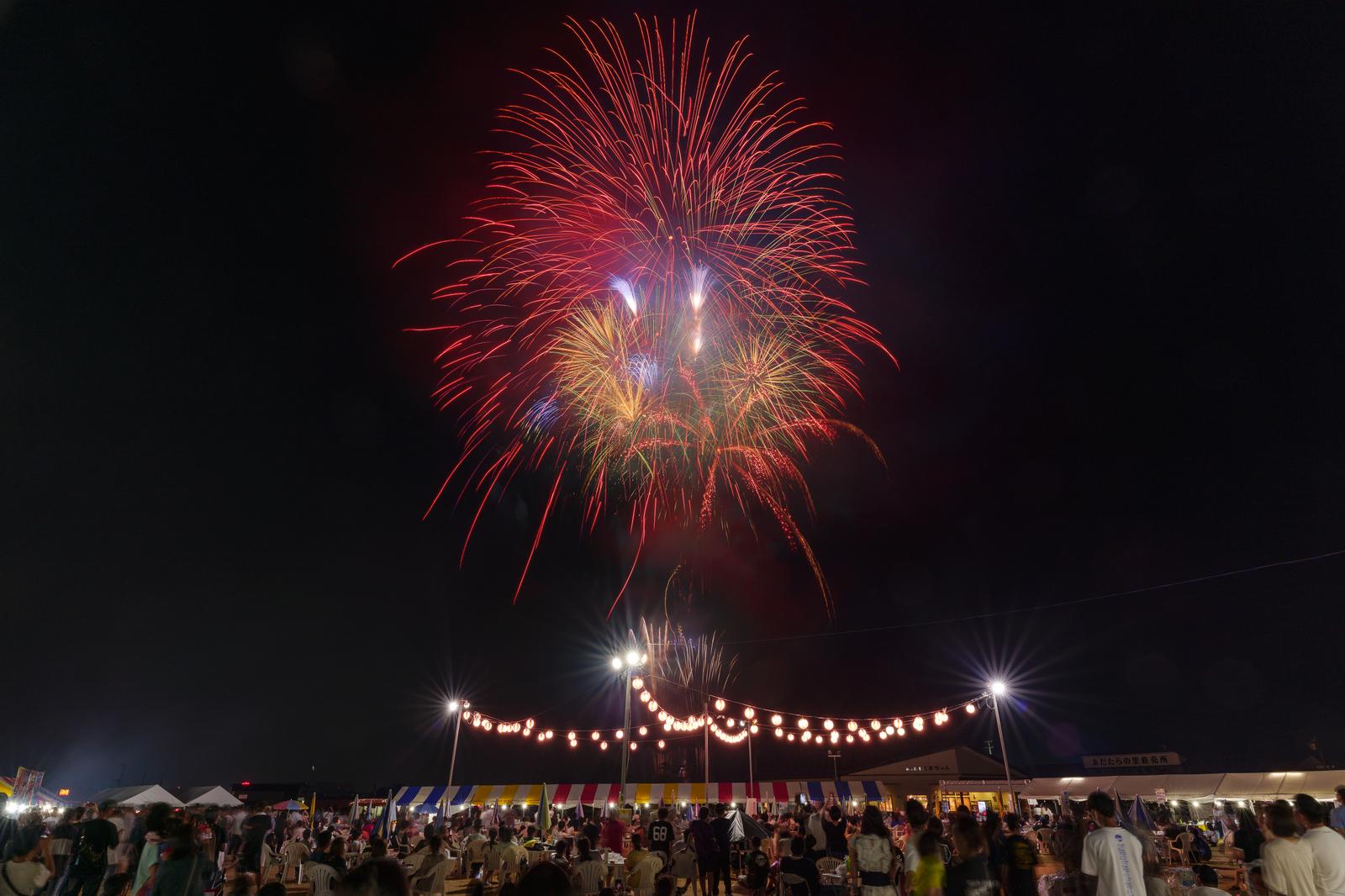 赤と金色の花火が夜空に打ち上がり、下方に屋台の灯りがある夏祭りの光景