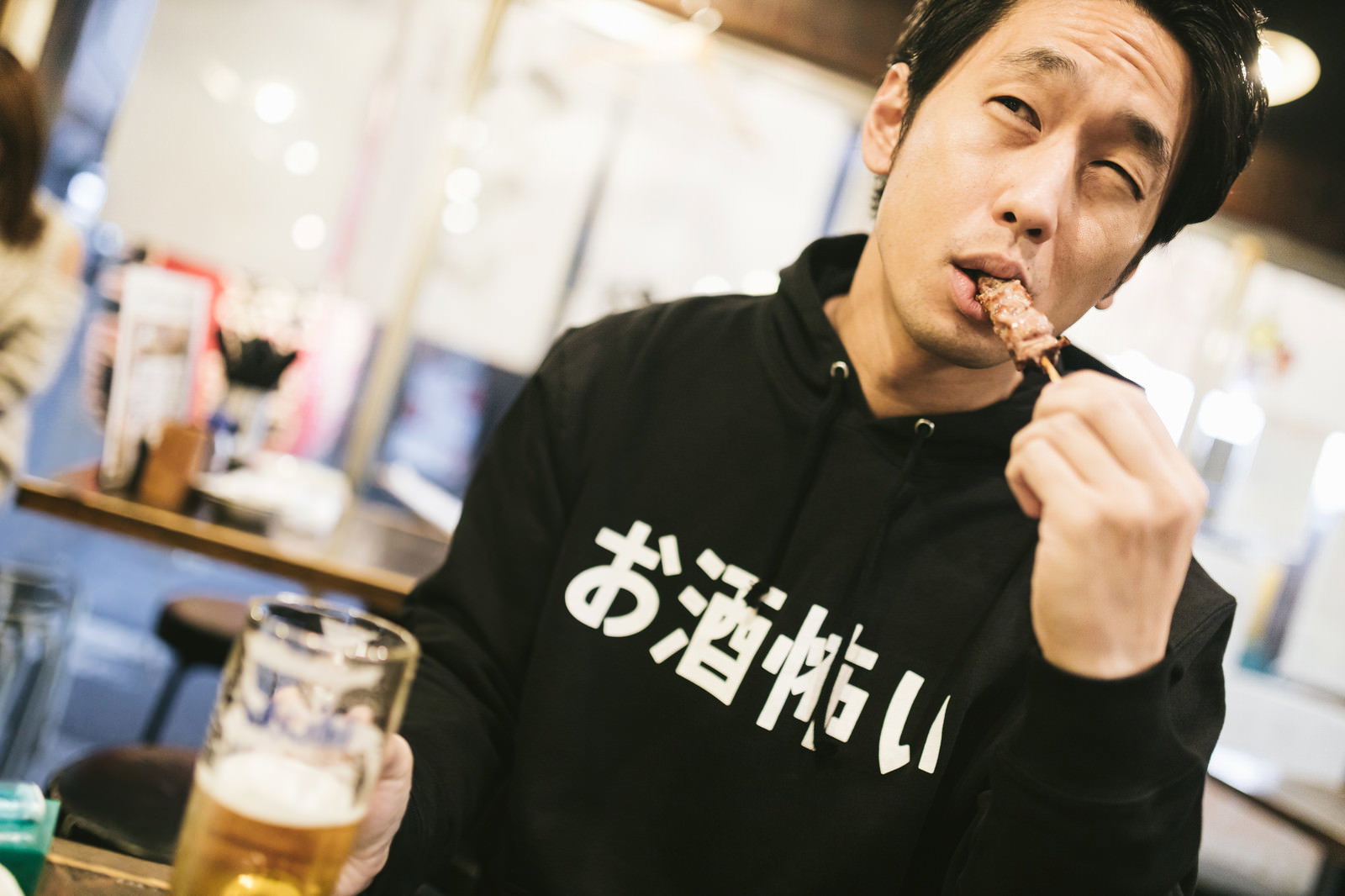 居酒屋でビールジョッキを持ち焼き鳥を食べる「お酒怖い」Tシャツの男性［モデル：大川竜弥］