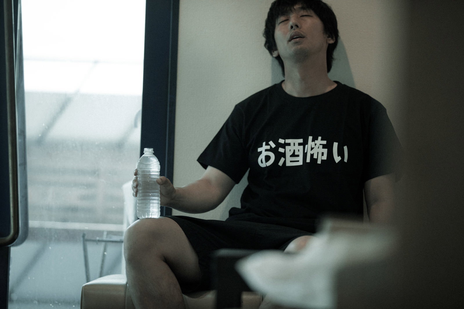 黒いTシャツを着た男性が室内の窓際でペットボトルを持ちながら座り込んでいる［モデル：大川竜弥］