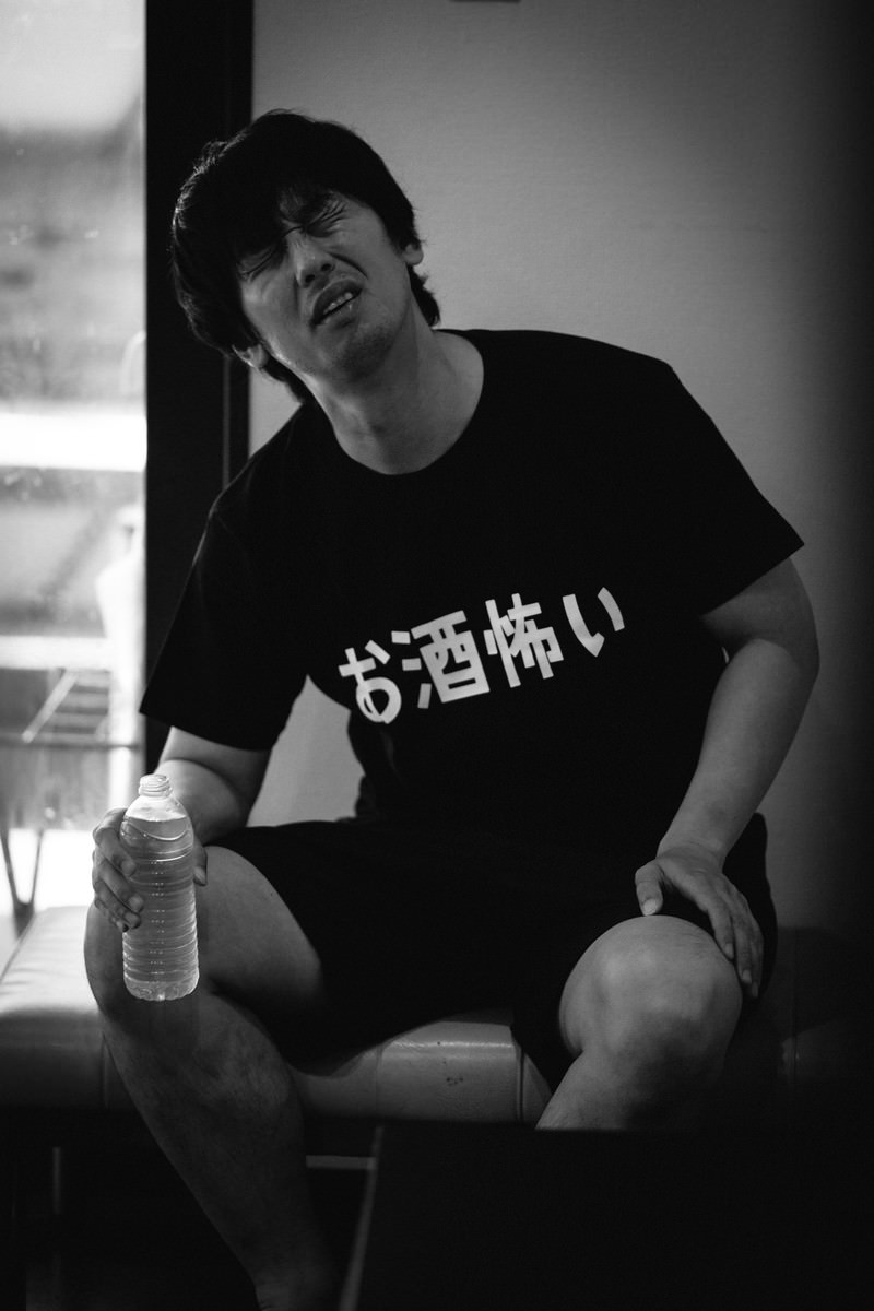 二日酔いで柱にもたれかかりながらペットボトルを持つ黒いTシャツの男性［モデル：大川竜弥］