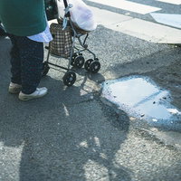 歩道に伸びる手押し車の影と水たまり、高齢者の買い物風景の写真