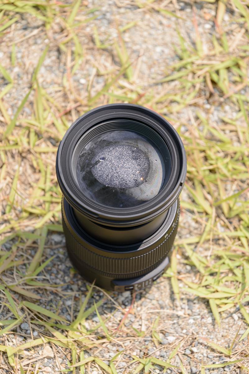 水没して内部に水滴が入ったズームレンズが草の上に置かれている