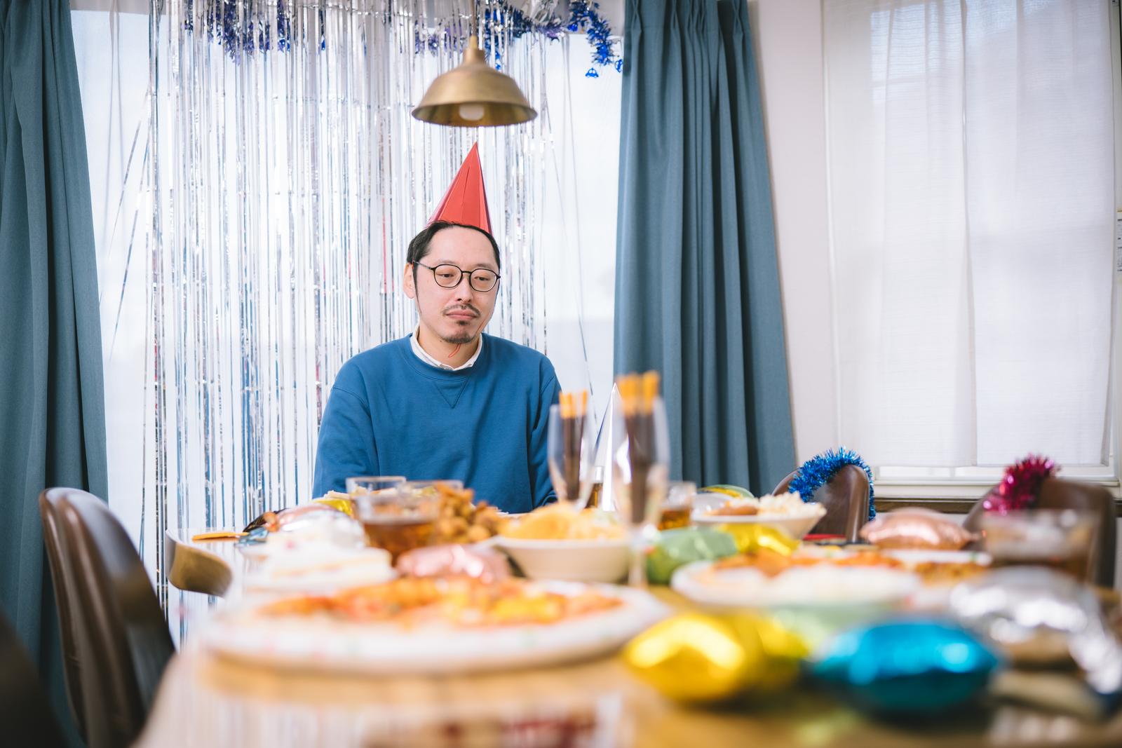 クリスマスパーティーの食卓で孤独そうに座る眼鏡の男性［モデル：ゆうせい］