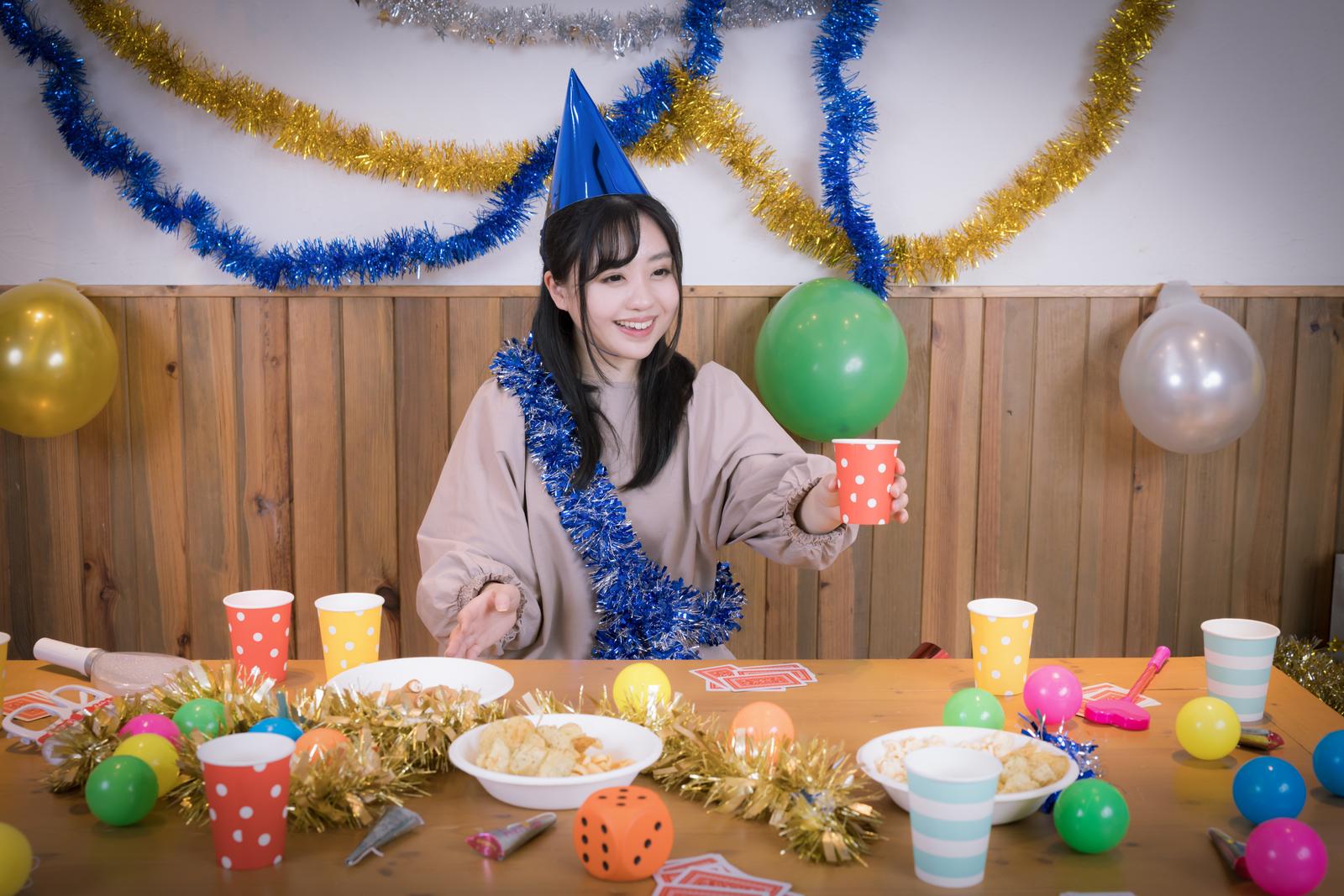 パーティーハットを被った女性が紙コップを持ち、テーブルで一人乾杯している［モデル：河村友歌］