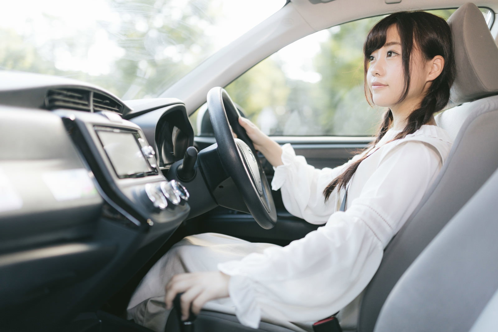 白いシャツを着た女性が車の運転席でハンドルを握っている［モデル：河村友歌］