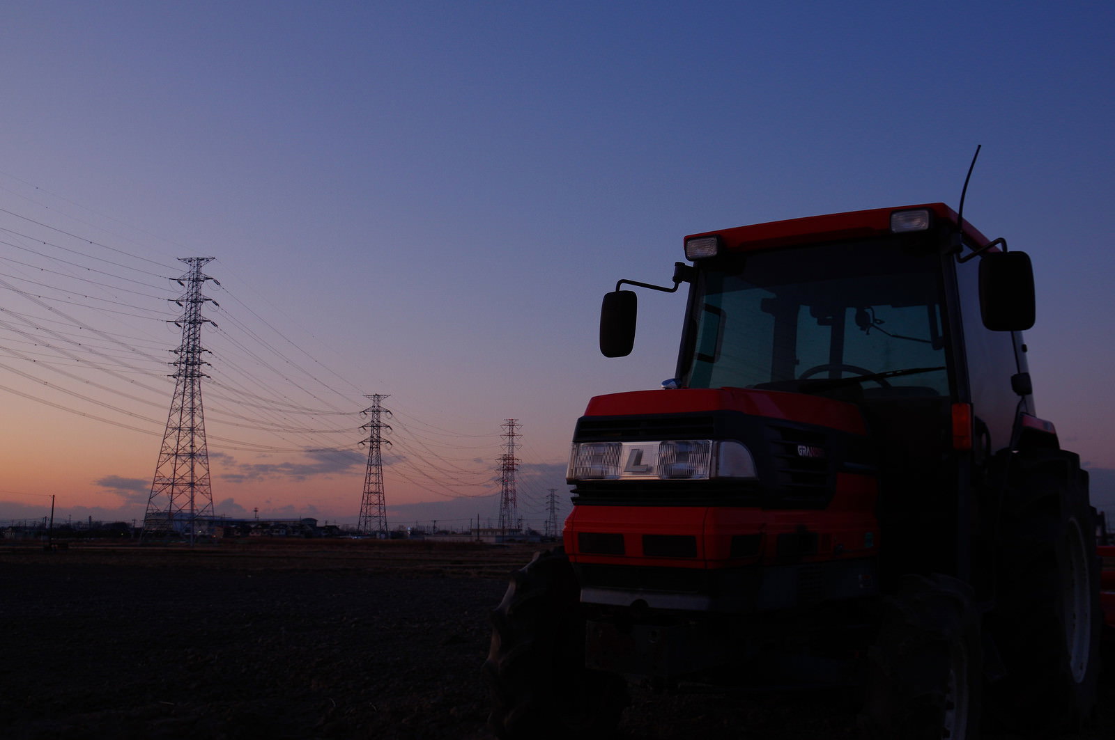 夕暮れの空を背景に送電線とトラクターのシルエットが映る田園風景
