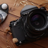 木目テーブルに置かれたペンタックスの一眼レフカメラとバケペン、露出計の写真