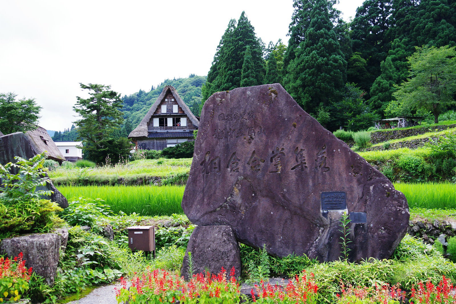 富山県南砺市の相倉集落で、緑の田畑に囲まれて立つ石碑と背景の合掌造り家屋