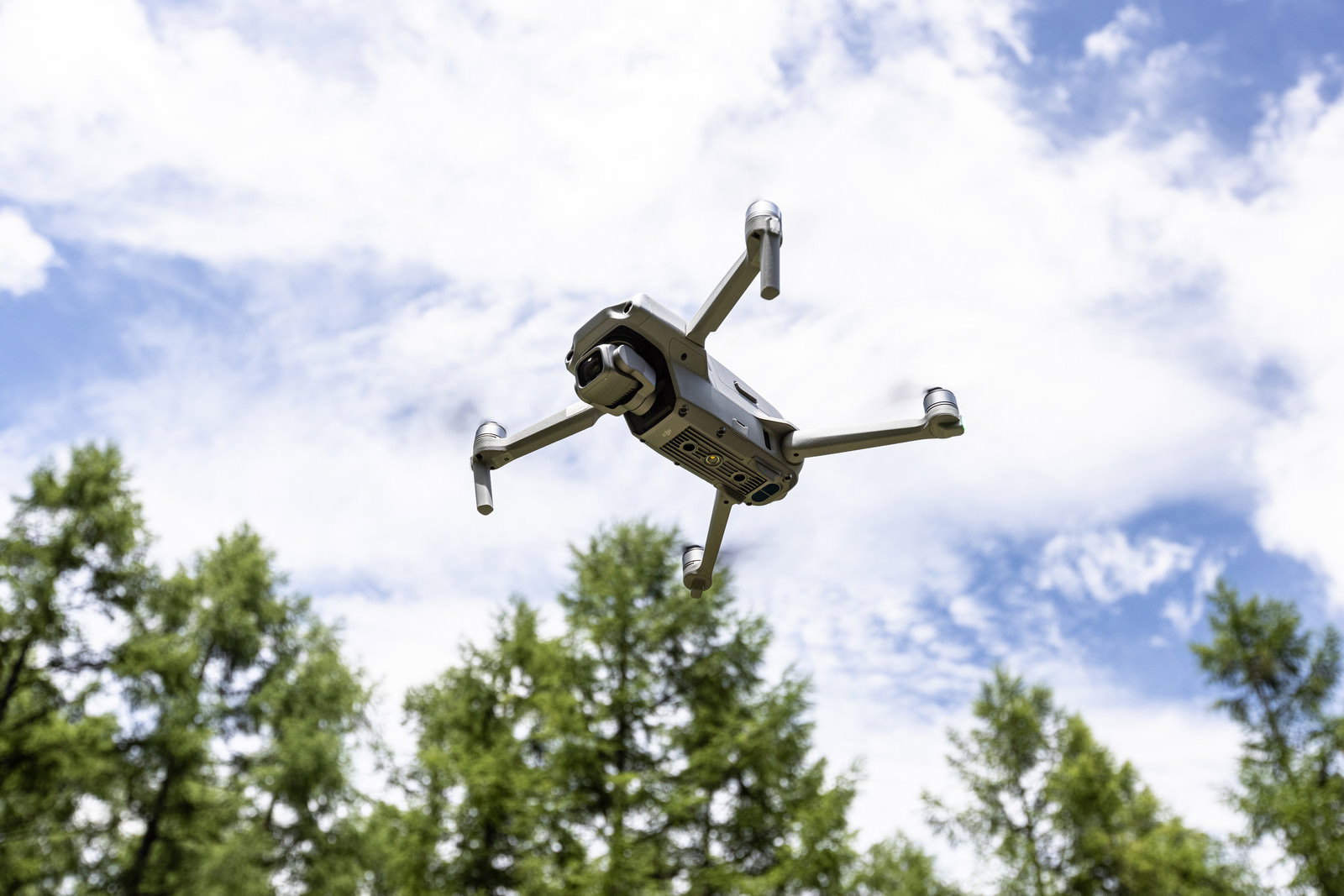 青空と雲を背景に緑の木々の上空でホバリングするDJIドローン