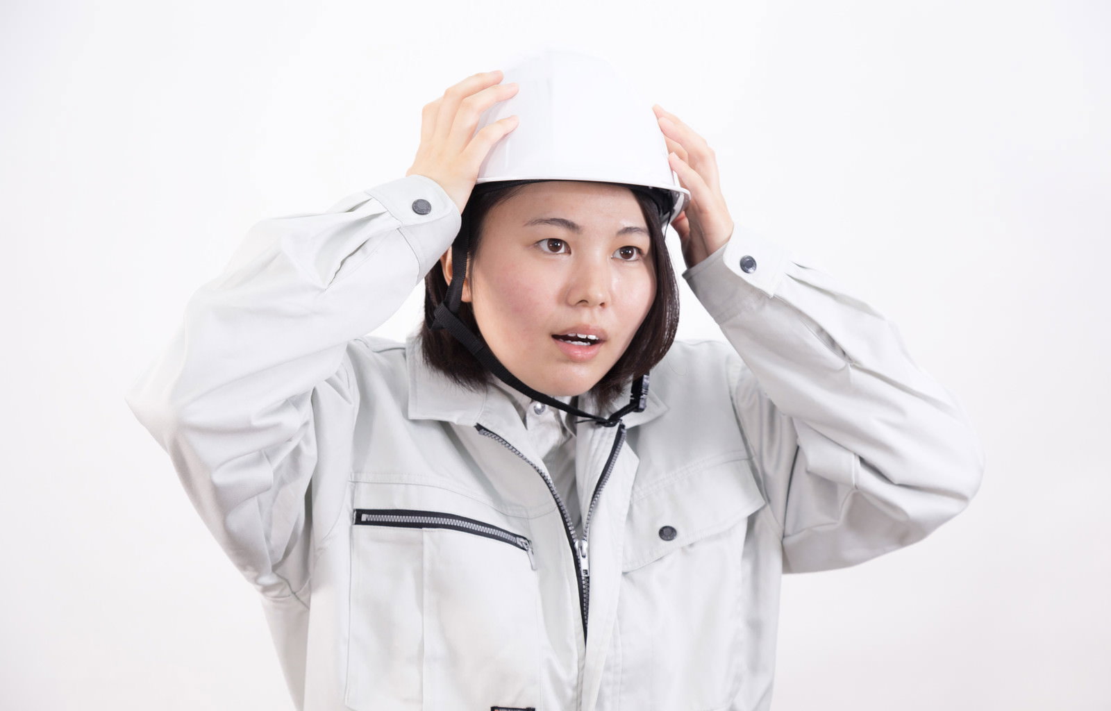 白いヘルメットと作業服を着用し、両手でヘルメットを押さえる女性作業員［モデル：佐竹PP］