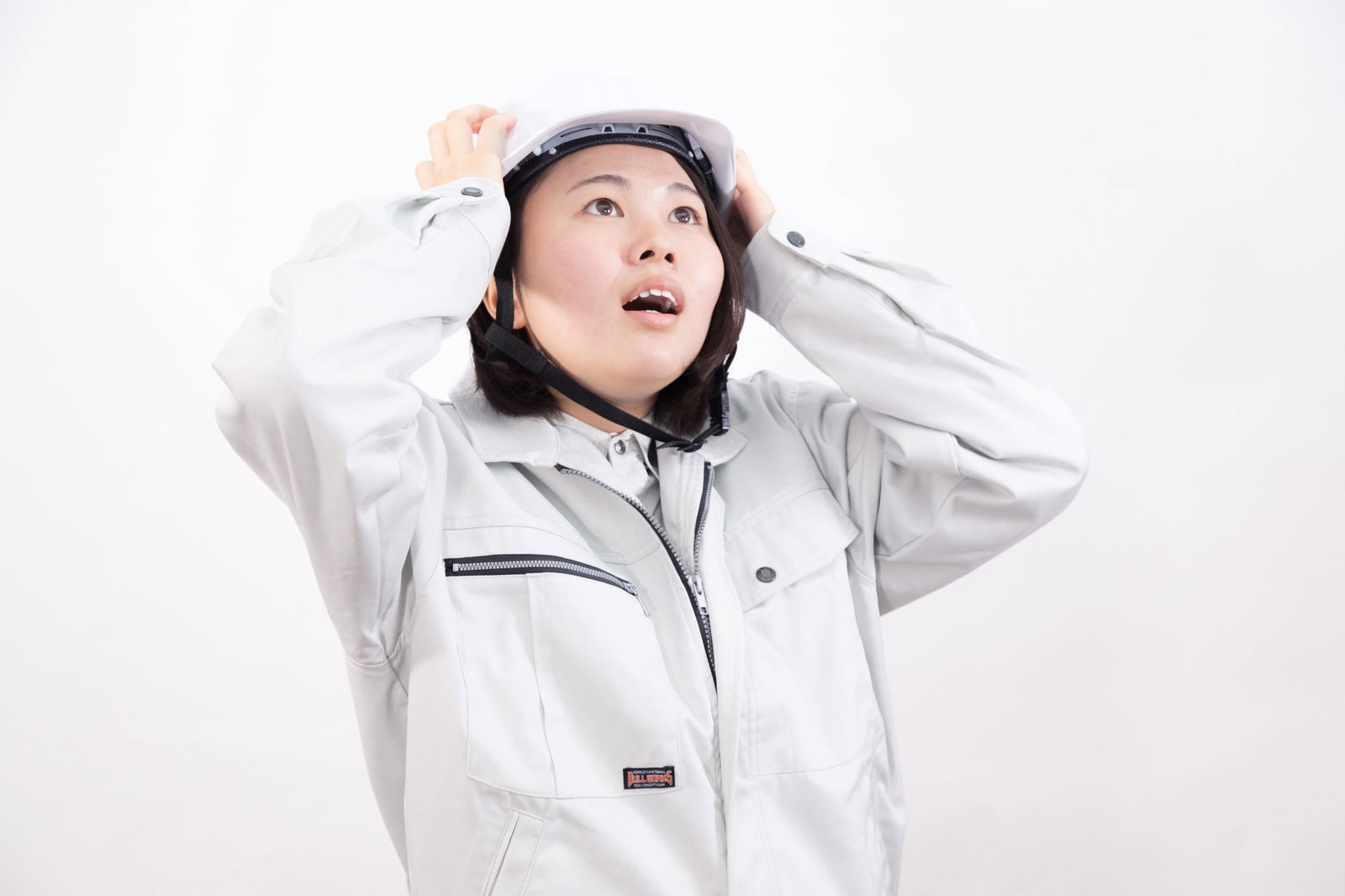 両手でヘルメットを押さえるグレーの作業服を着た女性作業員［モデル：佐竹PP］