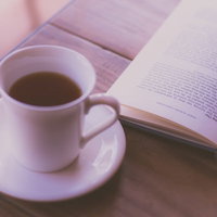 白いカップのコーヒーと開かれた洋書、木製テーブルの写真