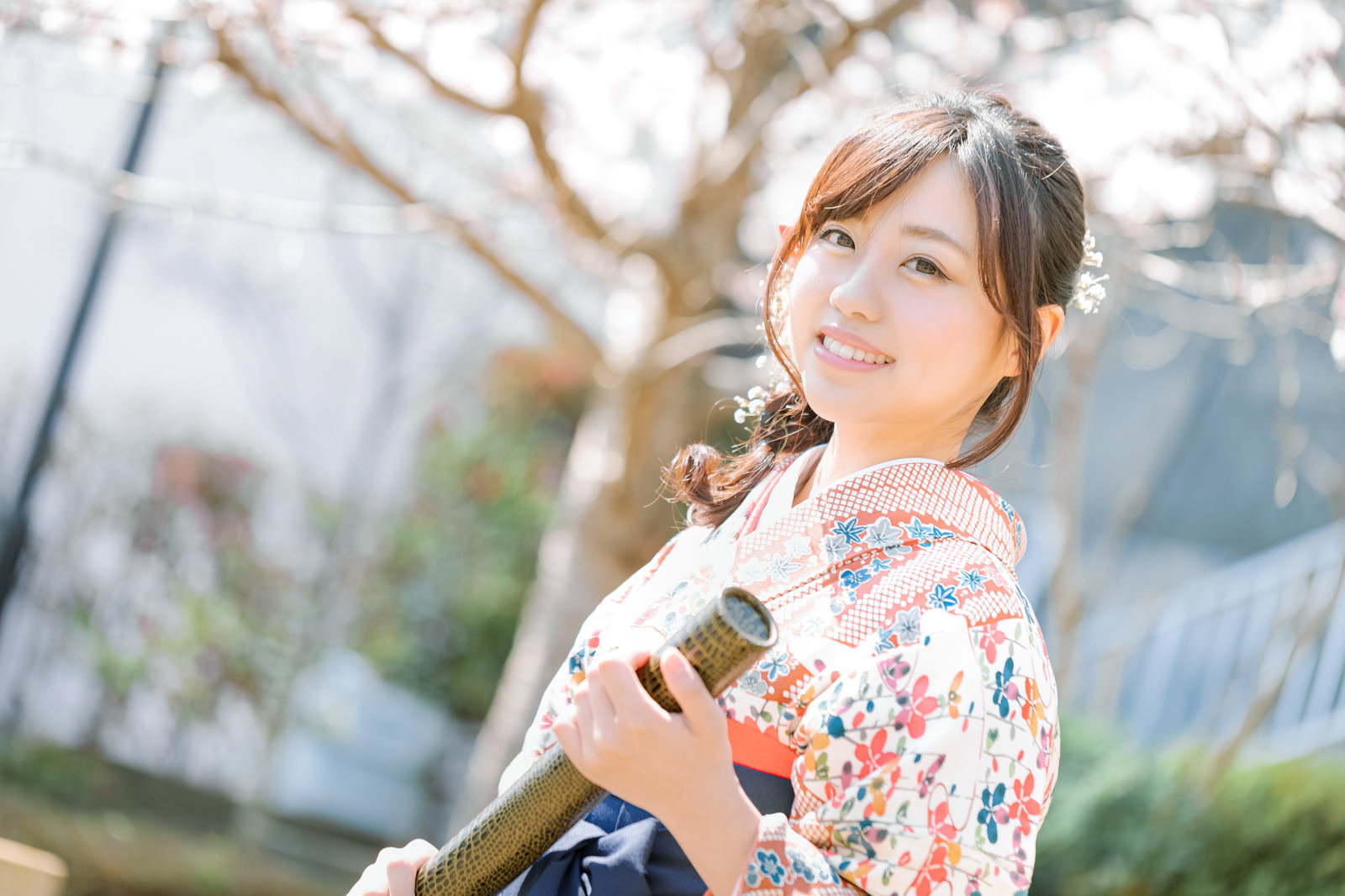 桜の木の下で卒業証書を持ち微笑む袴姿の女性［モデル：河村友歌］