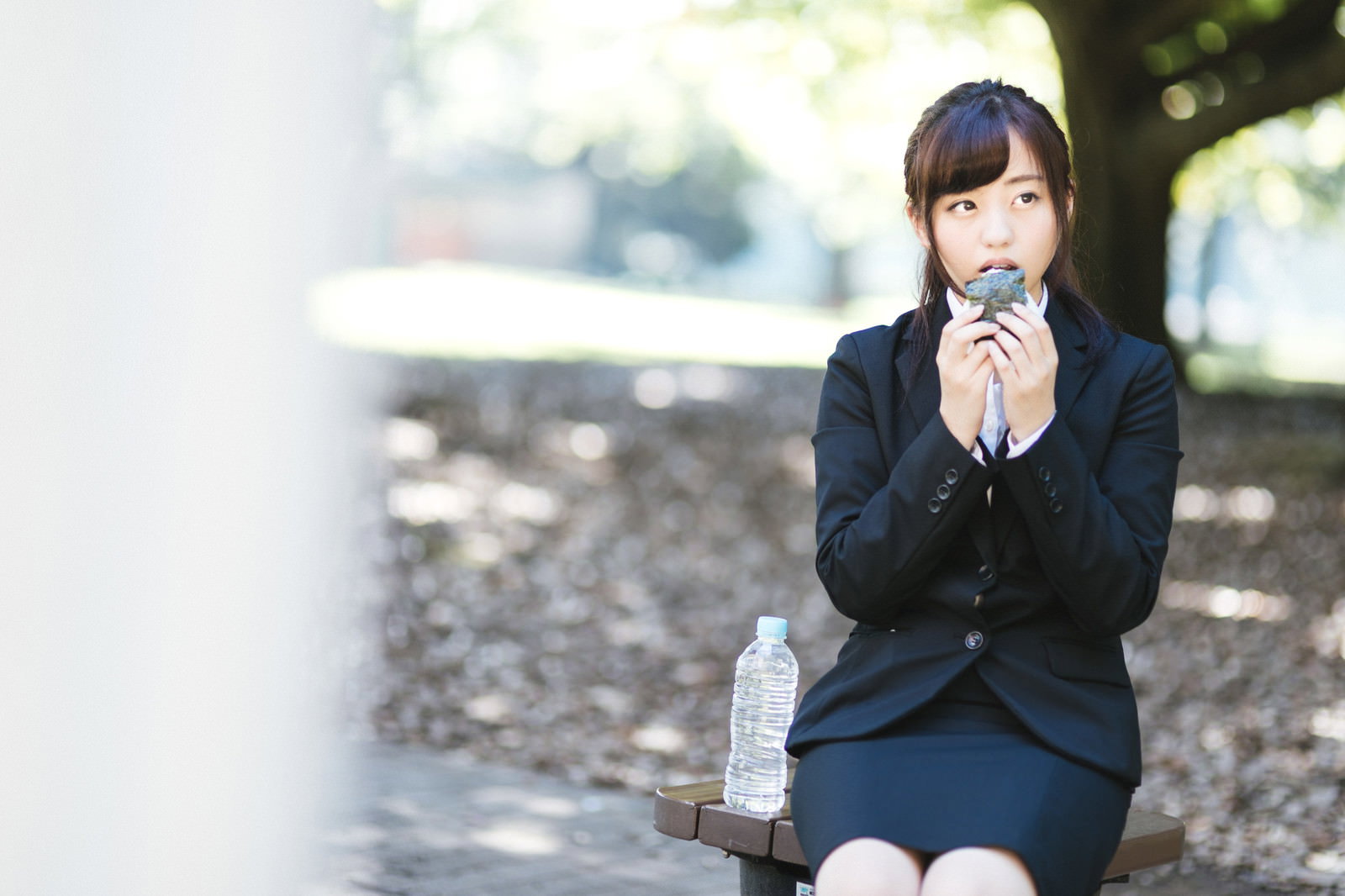 屋外のベンチで一人座っておにぎりを食べるスーツ姿の女性［モデル：河村友歌］