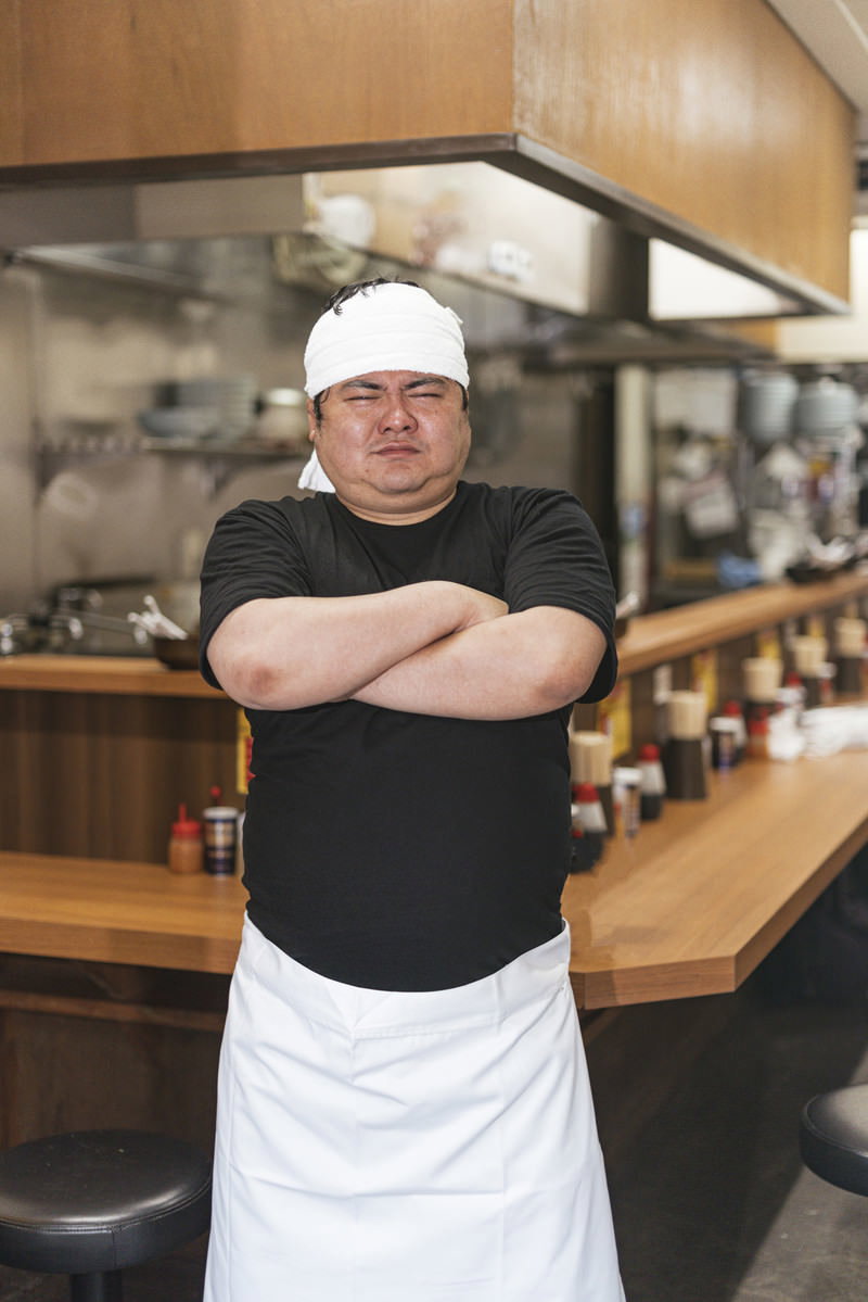 白いコック帽とエプロンを着用してキッチンで腕組みをするラーメン屋の男性店主［モデル：段田隼人］