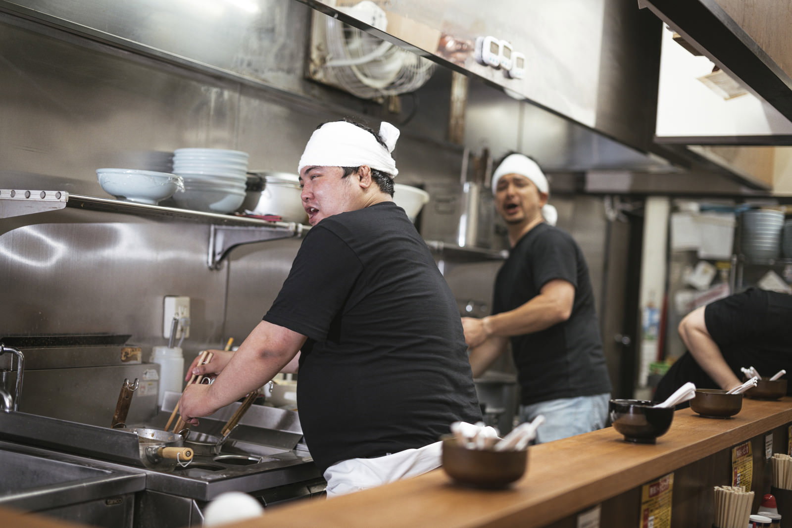 ラーメン店の厨房で白い帽子とエプロンを着けて調理作業をする男性店員。営業中の厨房の様子［モデル：段田隼人 Max_Ezaki 大川竜弥］