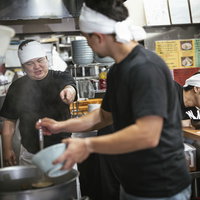 厨房でアルバイトに調理指導するラーメン屋の大将の写真