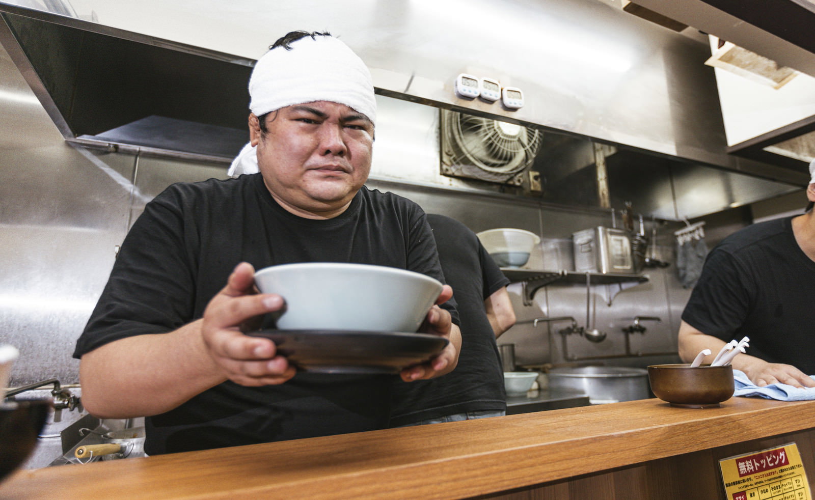 ラーメン店の厨房で緊張した表情でどんぶりを手に持つ白いコック帽の男性店主［モデル：段田隼人］